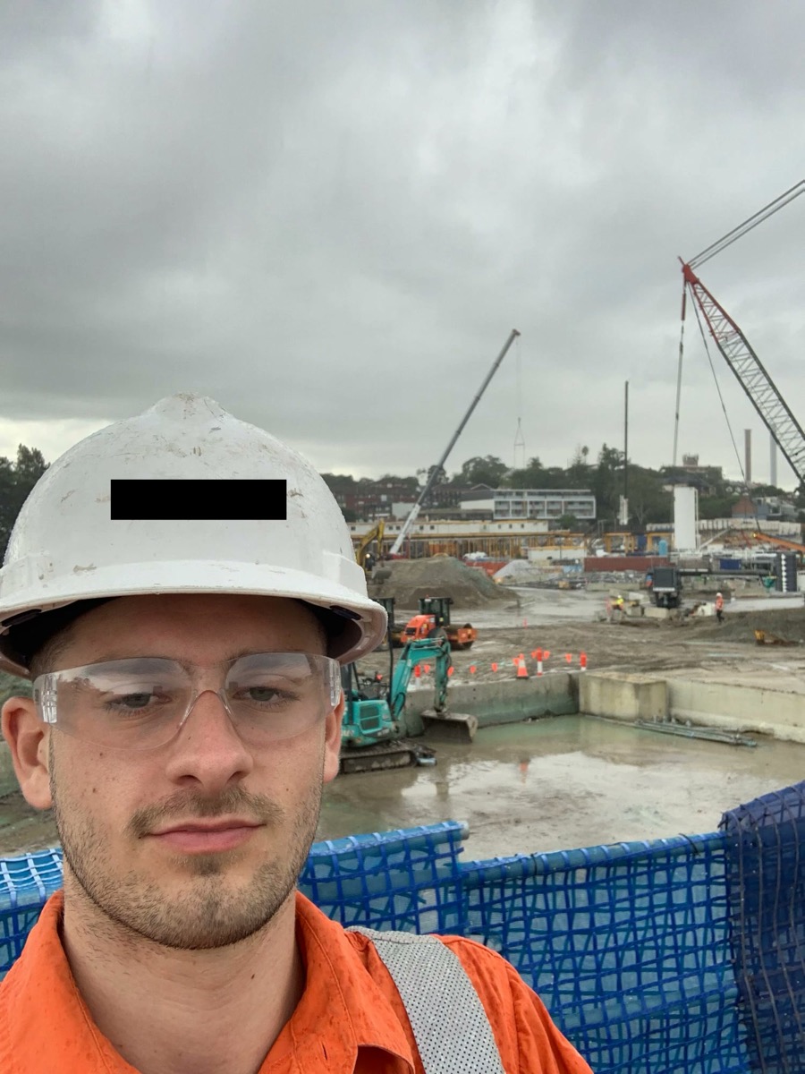 Adam in a hard hat and hi-vis on a construction site with cranes behind him